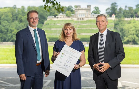 Drei Personen bei Unterzeichnung der Charta für Sterbende und Schwerstkranke vor Elbpanorama mit Schloss im Hintergrund.
