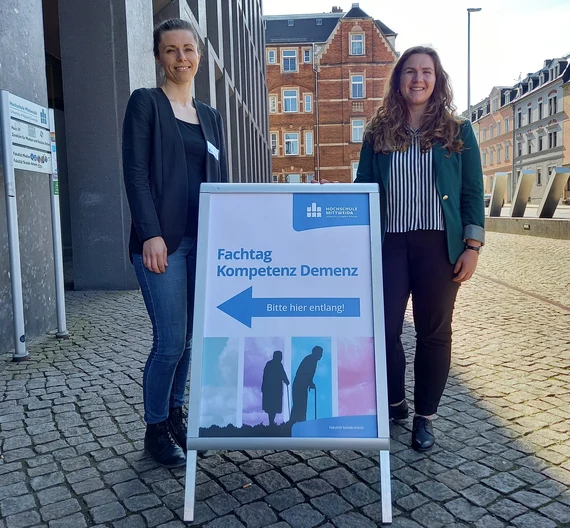 Zwei Frauen neben Aufsteller "Fachtag Kompetenz Demenz" der Hochschule Mittweida auf Platz vor Gebäude.