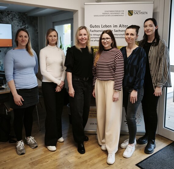 Die vier Kolleginnen der Fachservicestelle Sachsen stehen mit den zwei Kolleginnen des Kommunalen Sozialverbands vor einem Roll Up der Fachservicestelle.