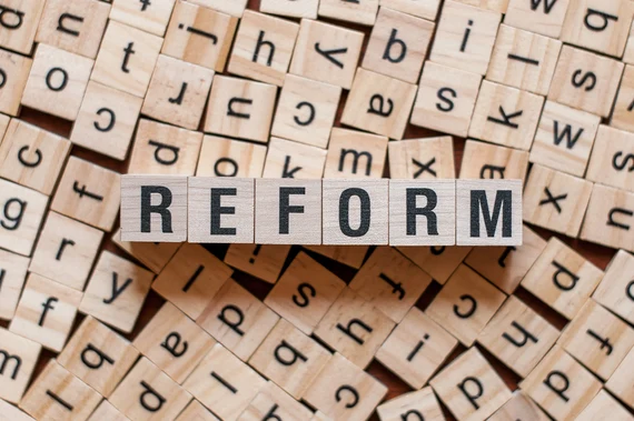 Holzbuchstaben bilden das Wort "REFORM" auf verstreuten Scrabble-Buchstaben.