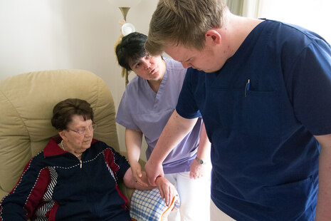 Ein Altenpflegeschüler fühlt bei einer Patientin den Puls. Angeleitet wird er dabei von seiner Ausbilderin.