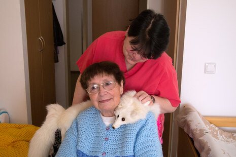 Eine ältere Dame hat einen Tierpelz um die Schulter gelegt. Ihre Pflegeperson schaut ihr liebevoll über die Schulter.