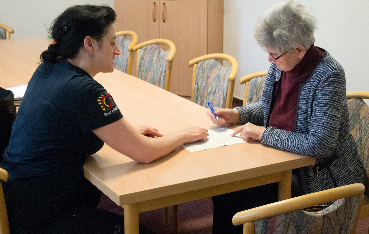 eine jüngere und ältere Frau sitzen an einem Tisch. Die jüngere Frau hilft beim Ausfüllen eines Dokuments. Der Link für zur Informationsseite »Beratung und Hilfe«