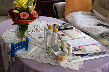 Ein Wohnzimmer-Tisch auf dem sind folgende Gegenstände zu sehen: Blumenvase mit Blumen, Getränke, Medikamente, eine Fernsehzeitung, ein Kalender, Fernbedienungen und eine Armbanduhr.