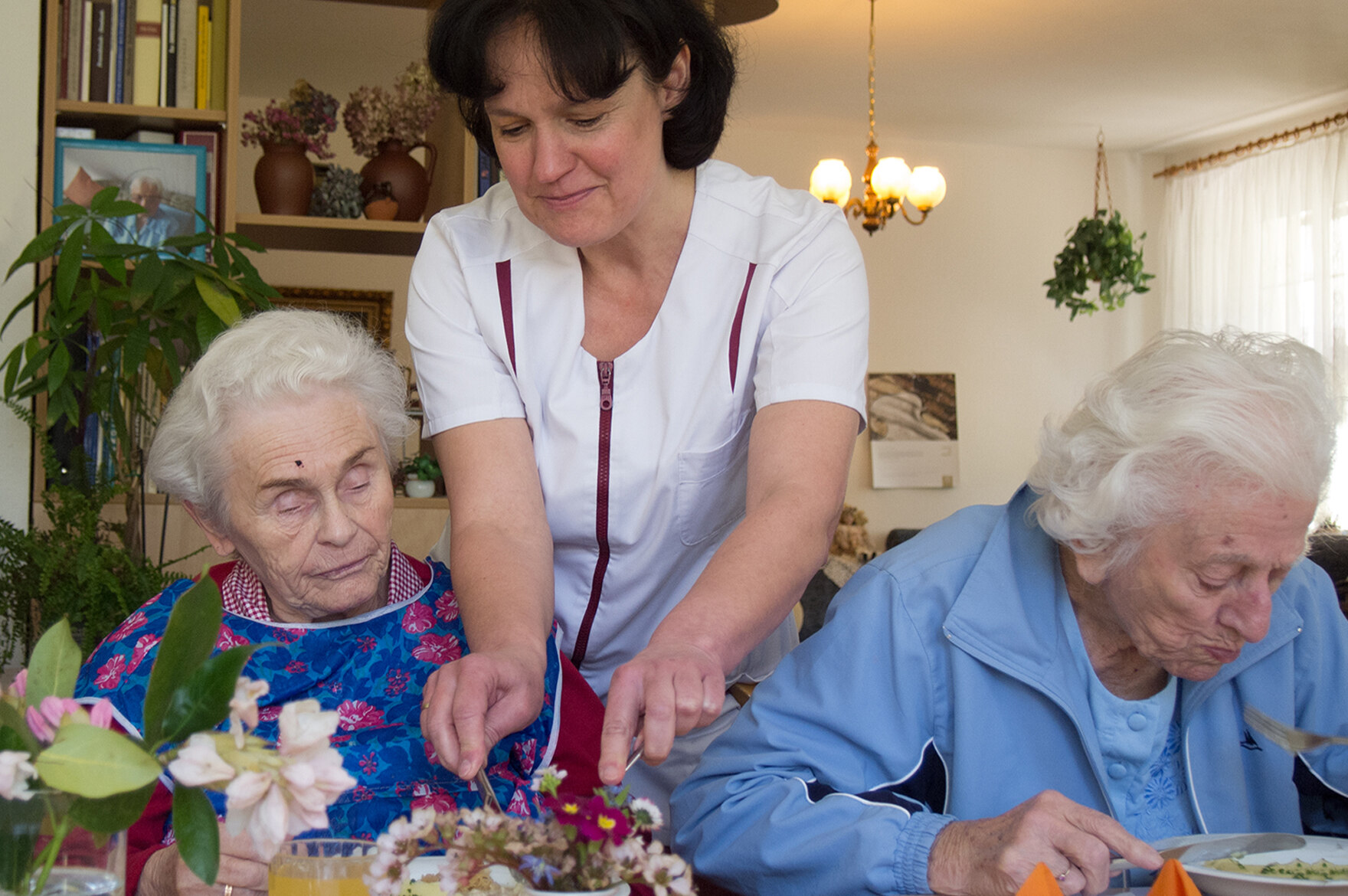 Zwei ältere Damen sitzen bei Essen. Eine Mitarbeiterin hilft beim Schneiden.