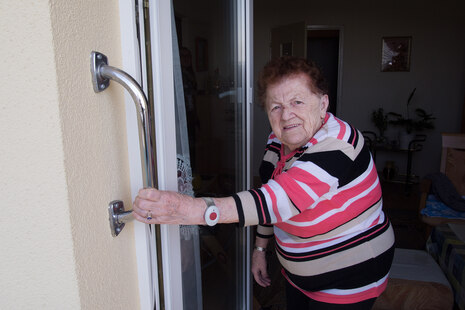 Ältere Dame nutzt einen Haltegriff, um sicher auf den Balkon zu steigen. Sie hat einen Notrufknopf ums Handgelenk.