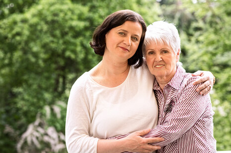 Mutter und Tochter umarmen sich und lächeln in die Kamera. Sie stehen in einem Garten.