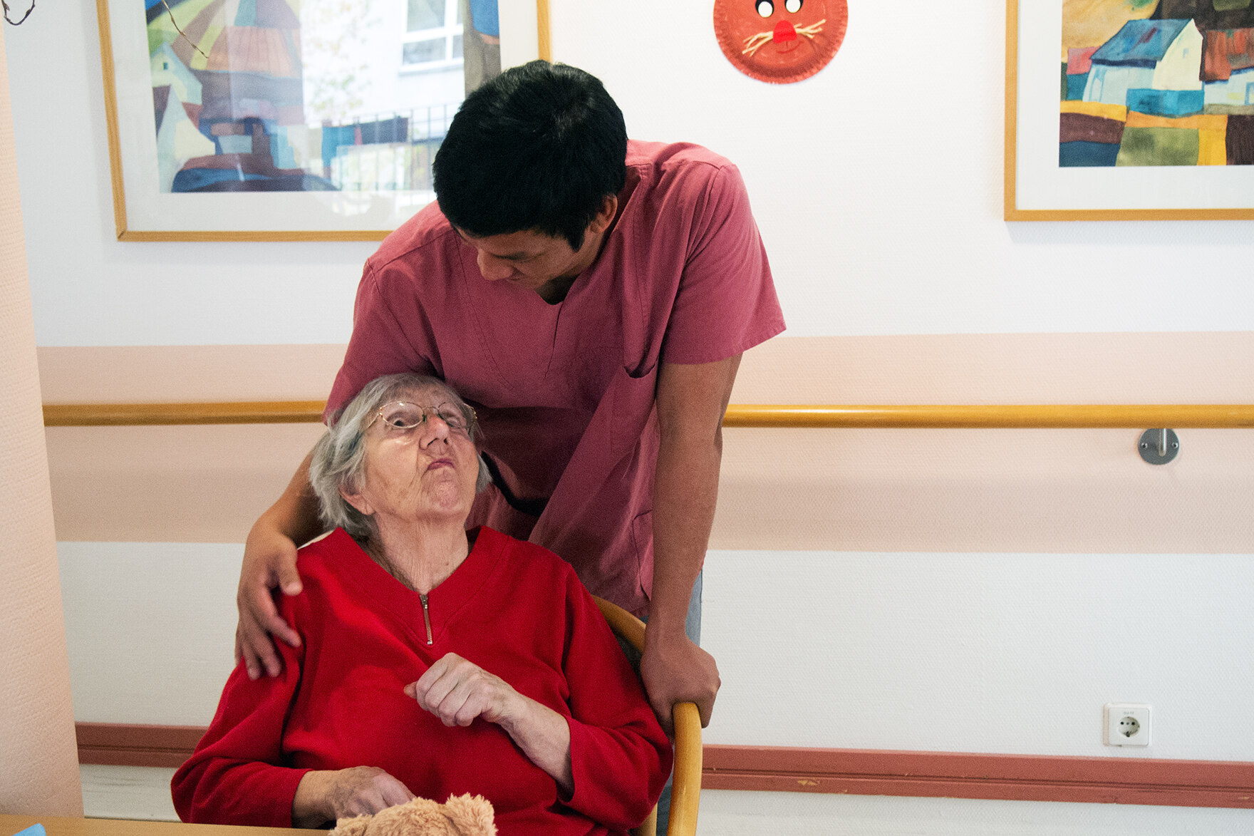 Ein Pfleger legt vertraut seine Hand an die Schulter einer älteren Bewohnerin und spricht mit ihr. Sie schaut ihn nach oben hin an.