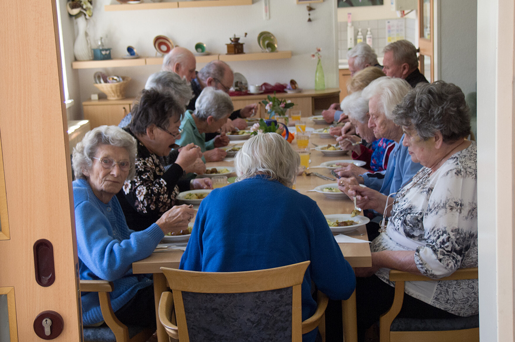 13 ältere Menschen essen gemeinsam an einem langen Tisch zum Mittag. Fotografiert wurde durch eine offene Schiebetür.