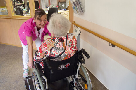 Eine Mitarbeiterin im Pflegeheim scherzt mit einer älteren Bewohnerin im Rollstuhl. Sie lacht.