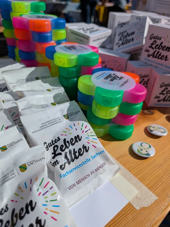 Auf diesen Bild sind die Goodies der Fachservicestelle Sachsen zu sehen. Vorne links befinden sich kleine Tütchen mit dem Logo der Fachservicestelle "Gutes Leben im Alter". Danach sind Textmarker und Tees zu finden.