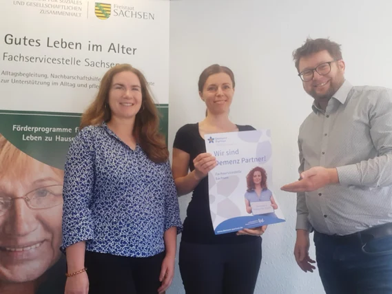 Drei Personen mit Plakat "Wir sind Demenz Partner!" vor Banner "Gutes Leben im Alter" der Fachservicestelle Sachsen.