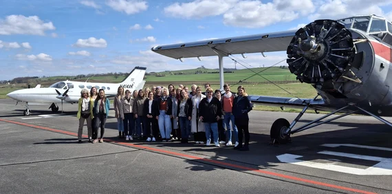 In dem Bild sieht man alle Teilnehmenden des 11. Nachbarschaftshelfer-Kontaktstellentreffen. Die Teilnehmenden stehen auf dem Flugplatz. Im Hintergrund sind zwei Flugzeuge zu sehen.