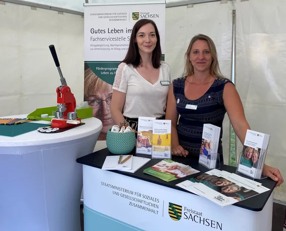 Zwei Mitarbeiterinnen am Infostand der Fachservicestelle Sachsen mit Broschüren und Wappen Freistaat Sachsen.