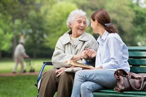 Seniorin im Rollstuhl und junge Betreuerin im Gespräch auf Parkbank, im Hintergrund Person auf Wiese.