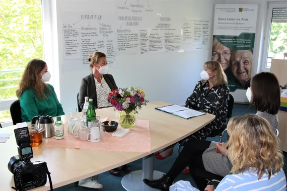 Fünf Personen mit Masken am Tisch bei Besprechung, Flipchart und Poster "Gutes Leben im Alter" im Hintergrund, Kamera vorn.