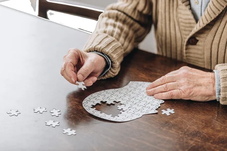 Hände einer Person in beigem Pullover beim Puzzeln; kreisförmiges Puzzle auf Holztisch mit fehlenden Teilen.